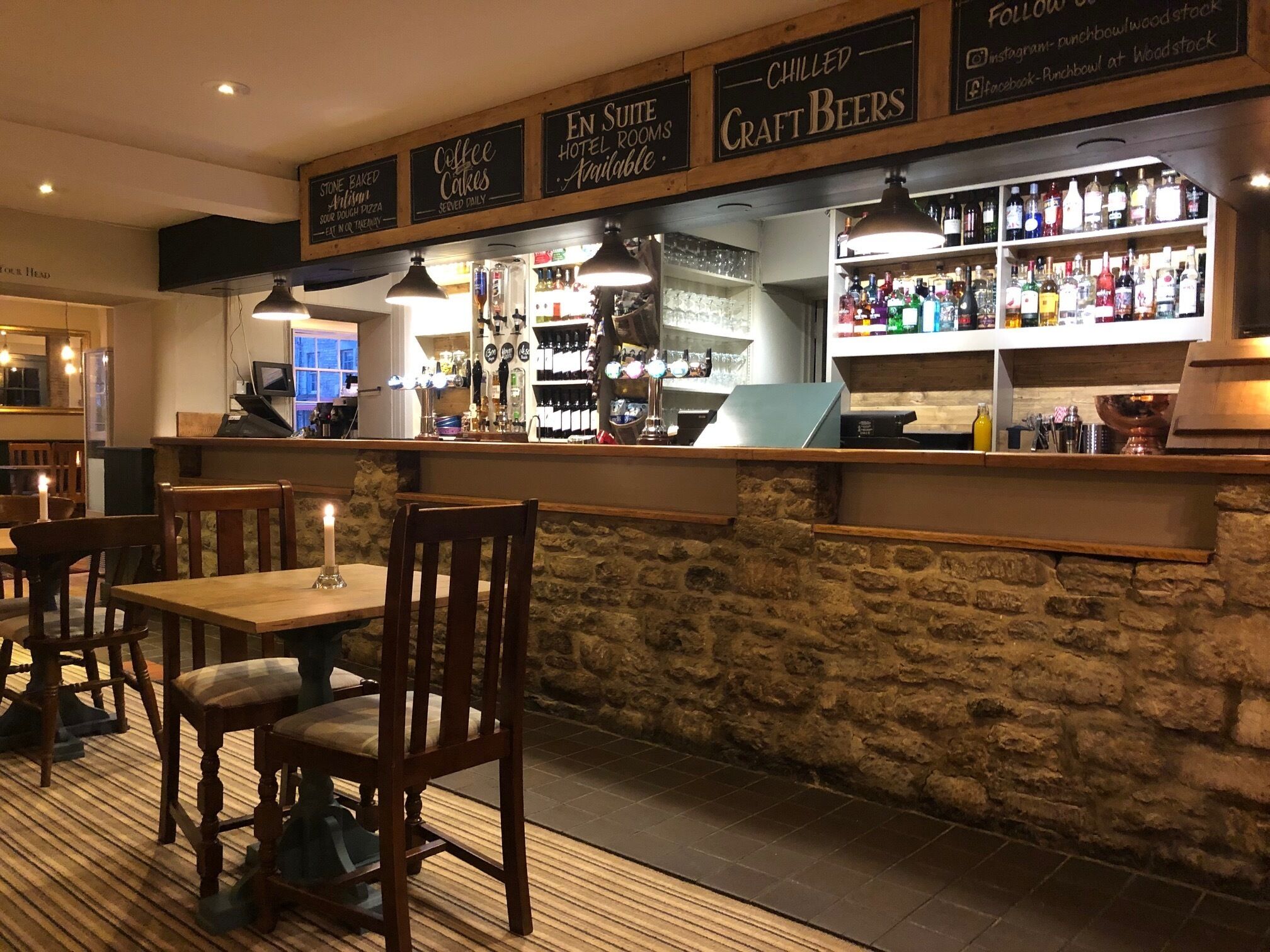 Eine Barbereich mit Steinwand, Holzsesseln und -tischen. Schilder weisen auf 'Coffee Cakes' und 'Chilled Craft Beers' hin.