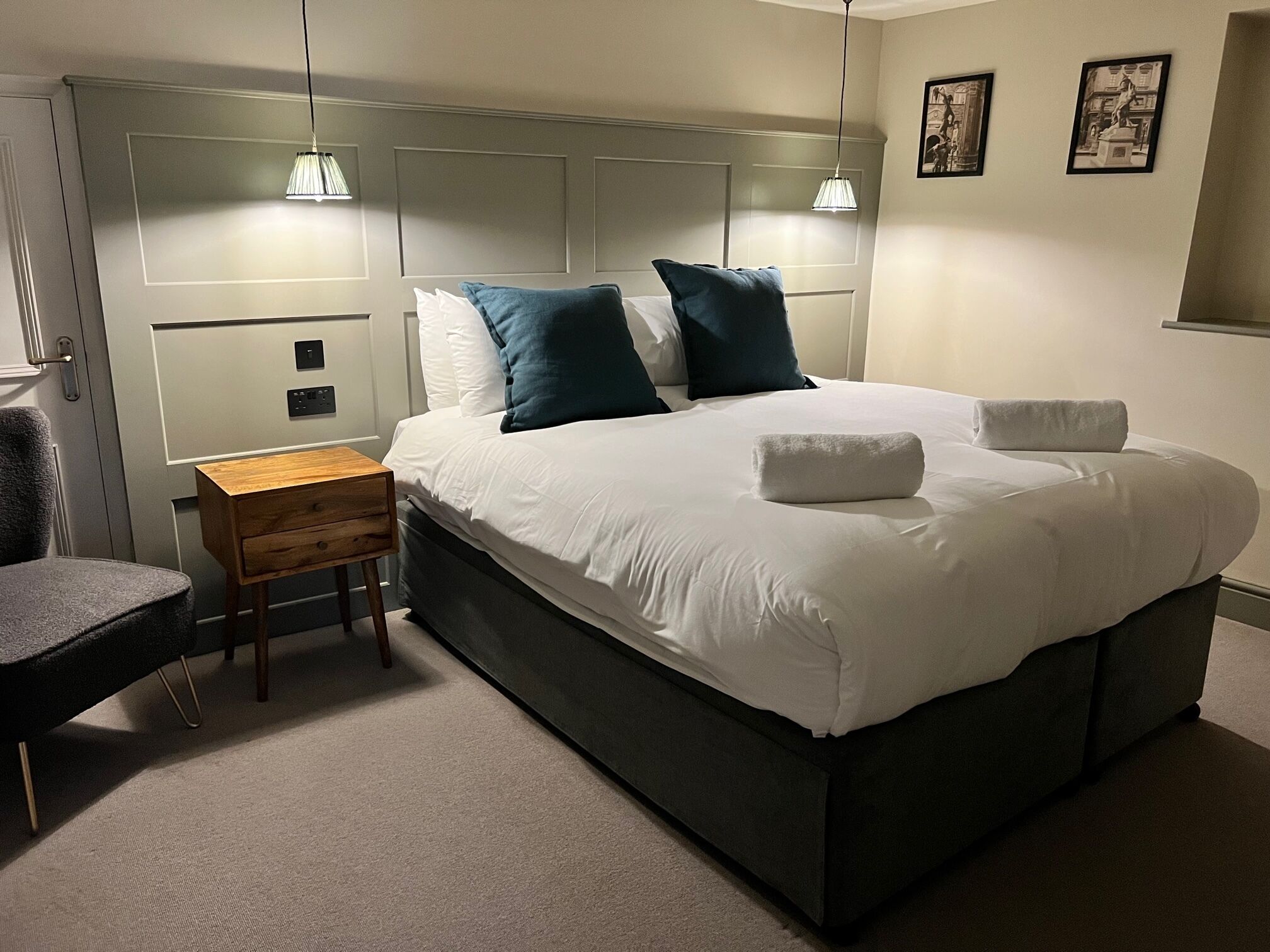 Un dormitorio ordenado con cama, mesita de noche, silla y lámparas de pared.