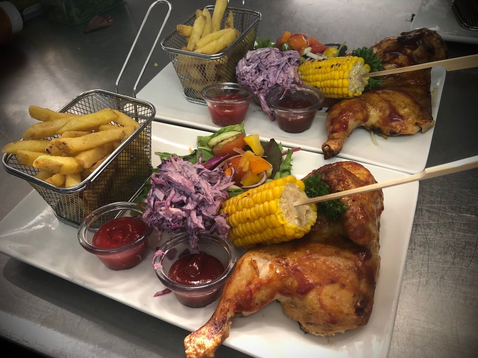 Un plato con un muslo de pollo asado, mazorca de maíz, ensalada de col morada y papas fritas.