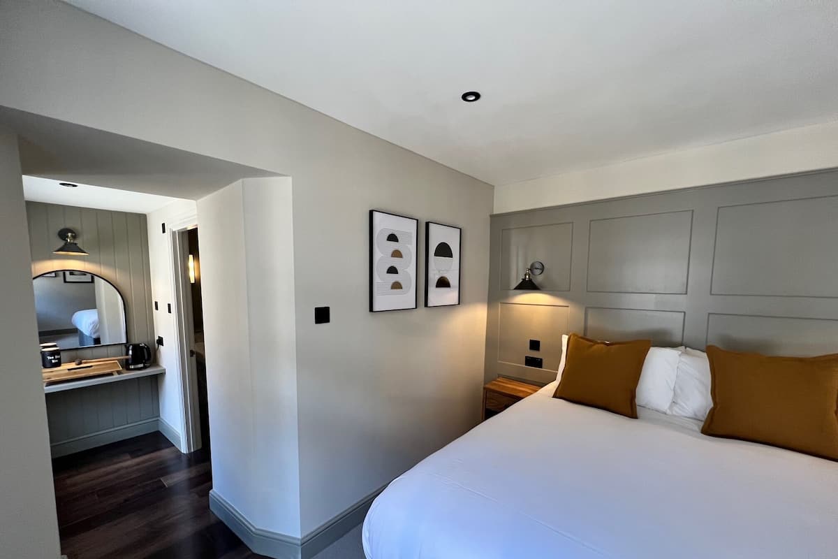 A modern hotel room with a large bed, a bedside table, a mirror, and a small desk with a lamp.