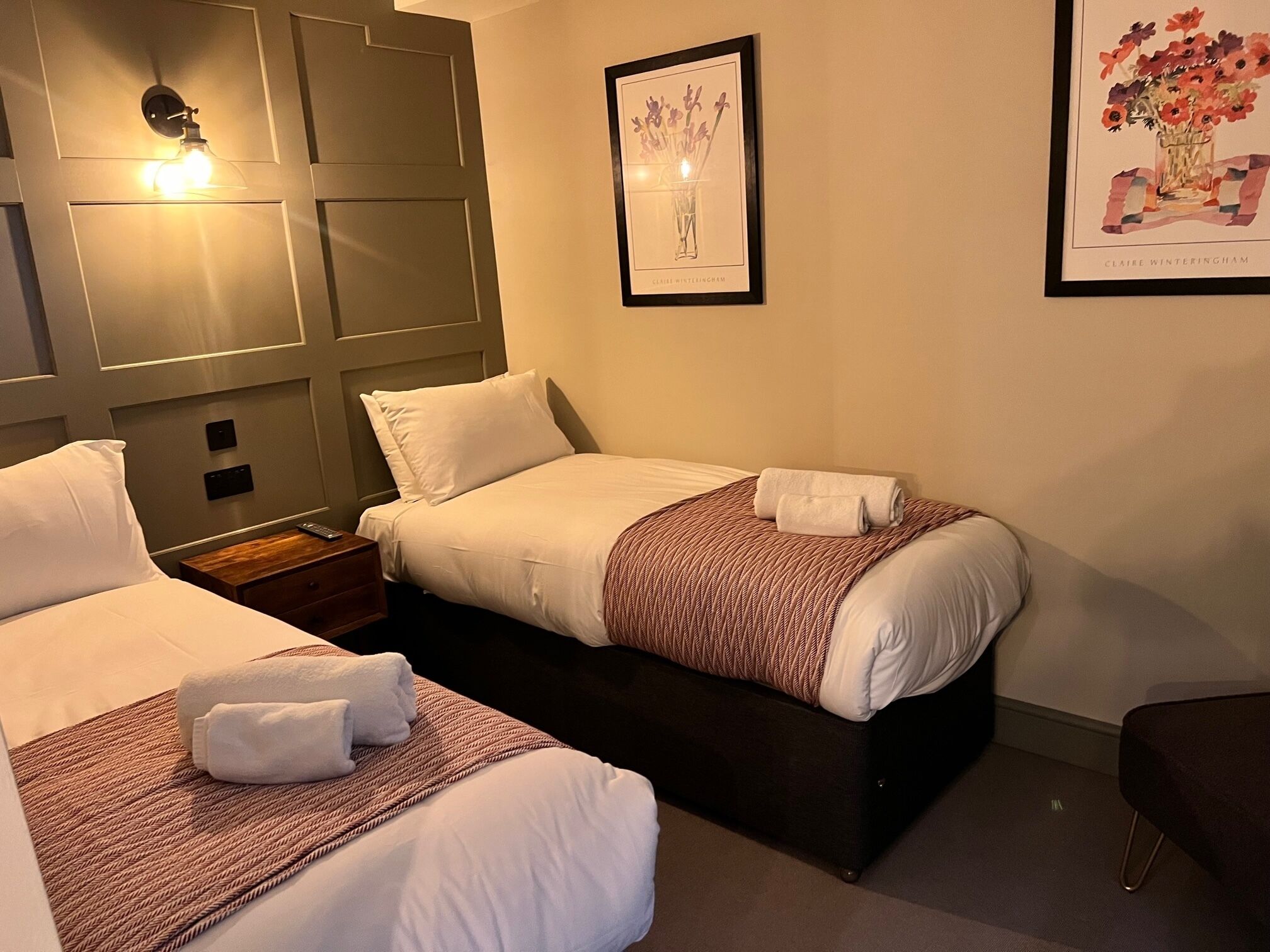 Habitación de hotel con dos camas, una mesita de noche, una lámpara de pared y dos obras de arte enmarcadas.