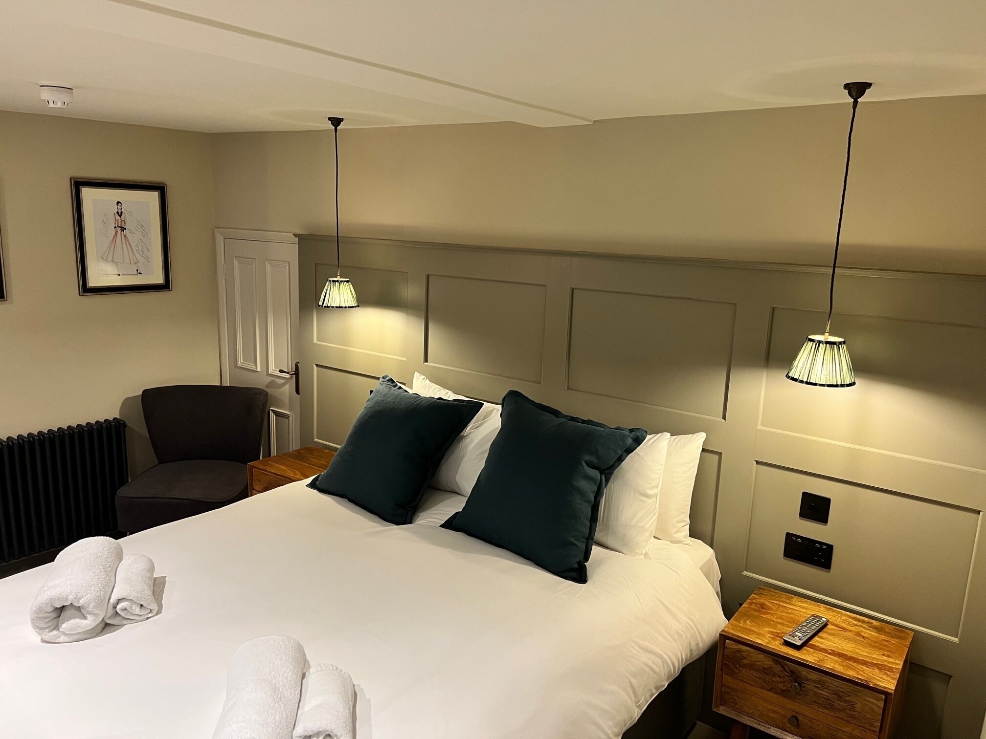 Una habitación de hotel con una cama grande, dos mesitas de noche, una silla, un radiador y un cuadro en la pared.