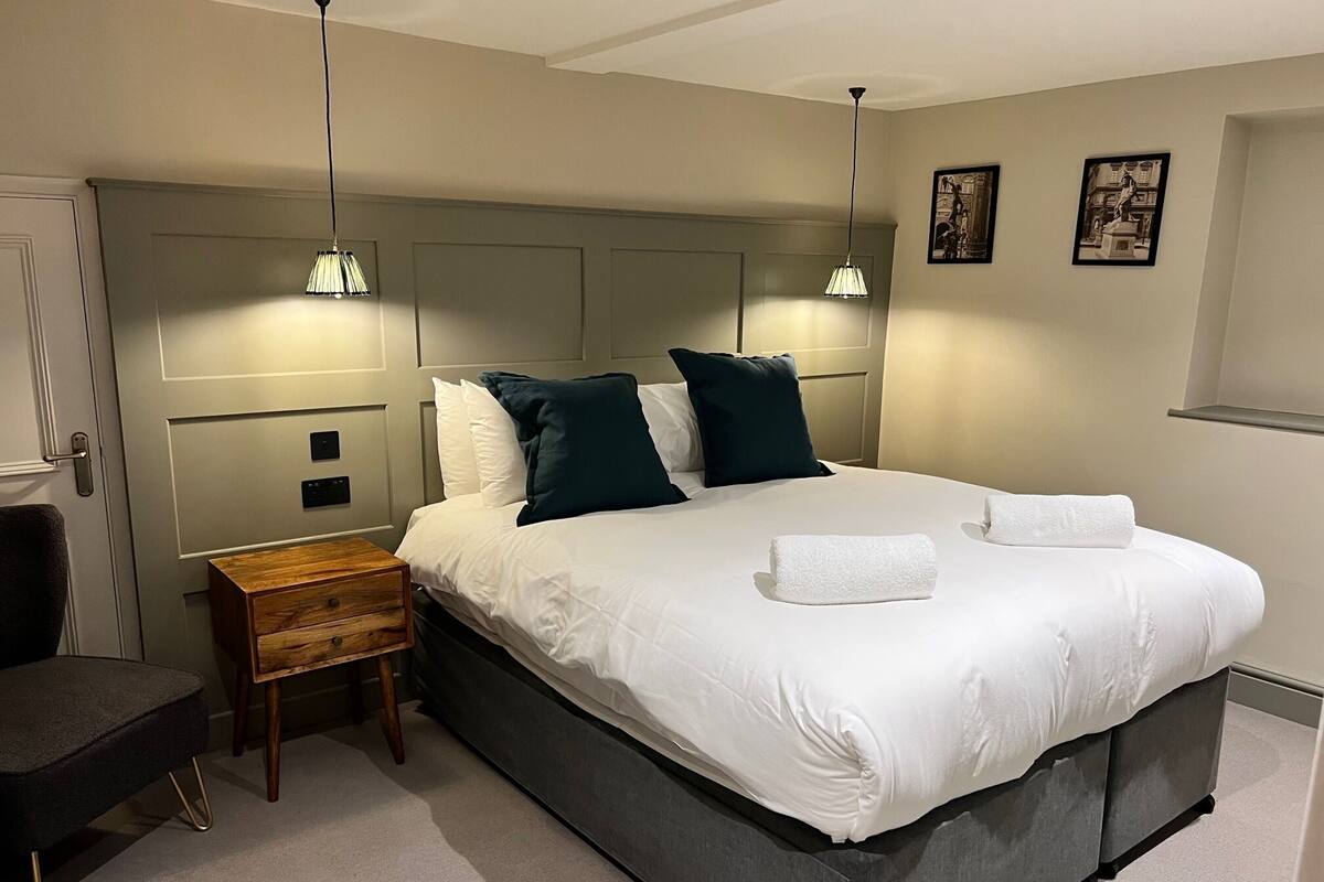 A neatly made bed with white linens and two pillows, a bedside table with a lamp, and a chair.