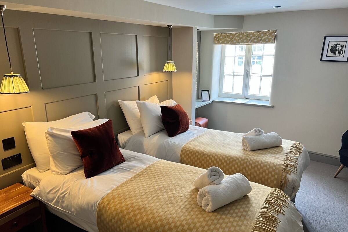A hotel room with two beds, each with a checkered bedspread and pillows. There are bedside lamps, a window with a curtain, and a framed picture on the wall.