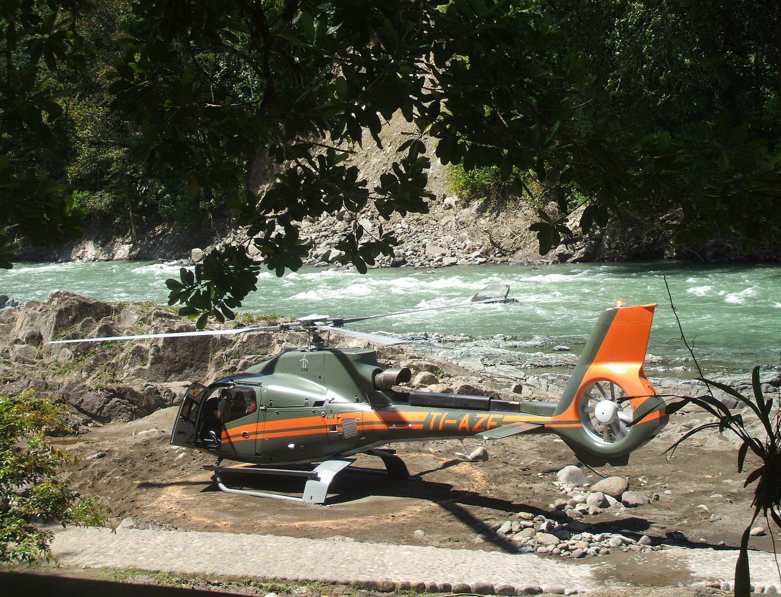 Un helicóptero estacionado cerca de un río con terreno rocoso y vegetación densa al fondo.