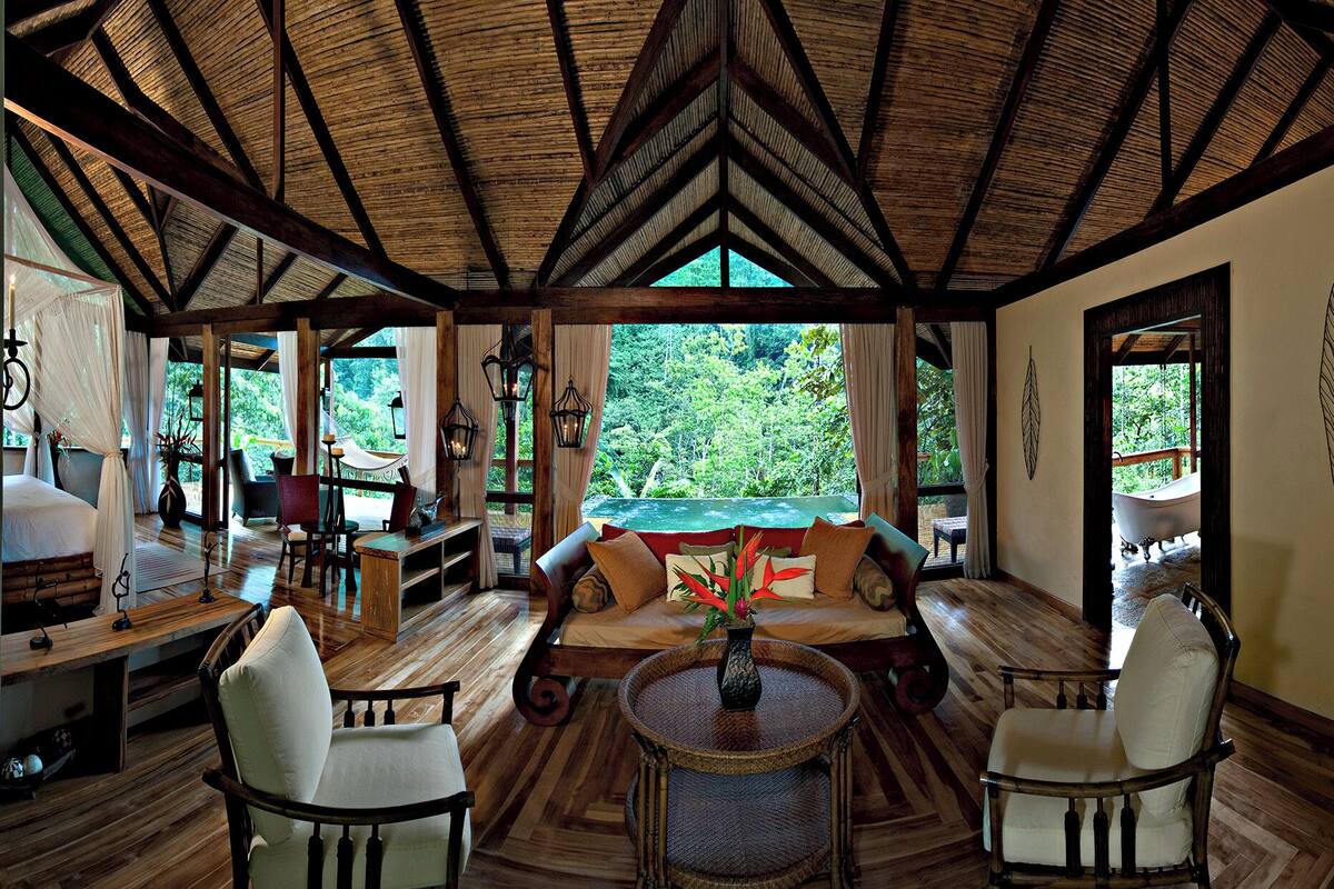 A spacious room with a high wooden ceiling, a large window offering a view of lush greenery, a comfortable seating area with a sofa and chairs, and a small round table with a vase.