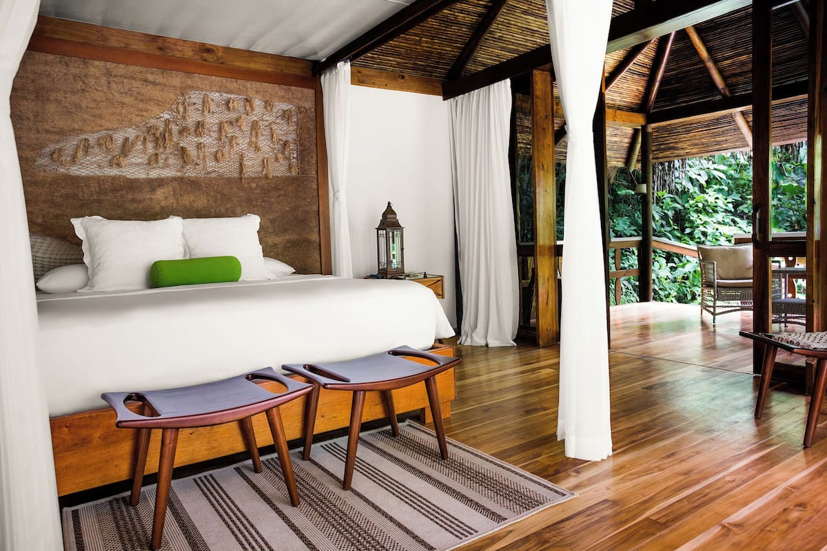 A bedroom with a bed, two chairs, a nightstand, a lantern, and a view of the outdoors.