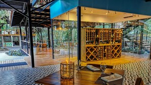 A restaurant with a modern design, featuring a wooden table and chairs, a glass wine rack, and a view of greenery outside. - Pacuare Lodge by Böëna (Siquirres)