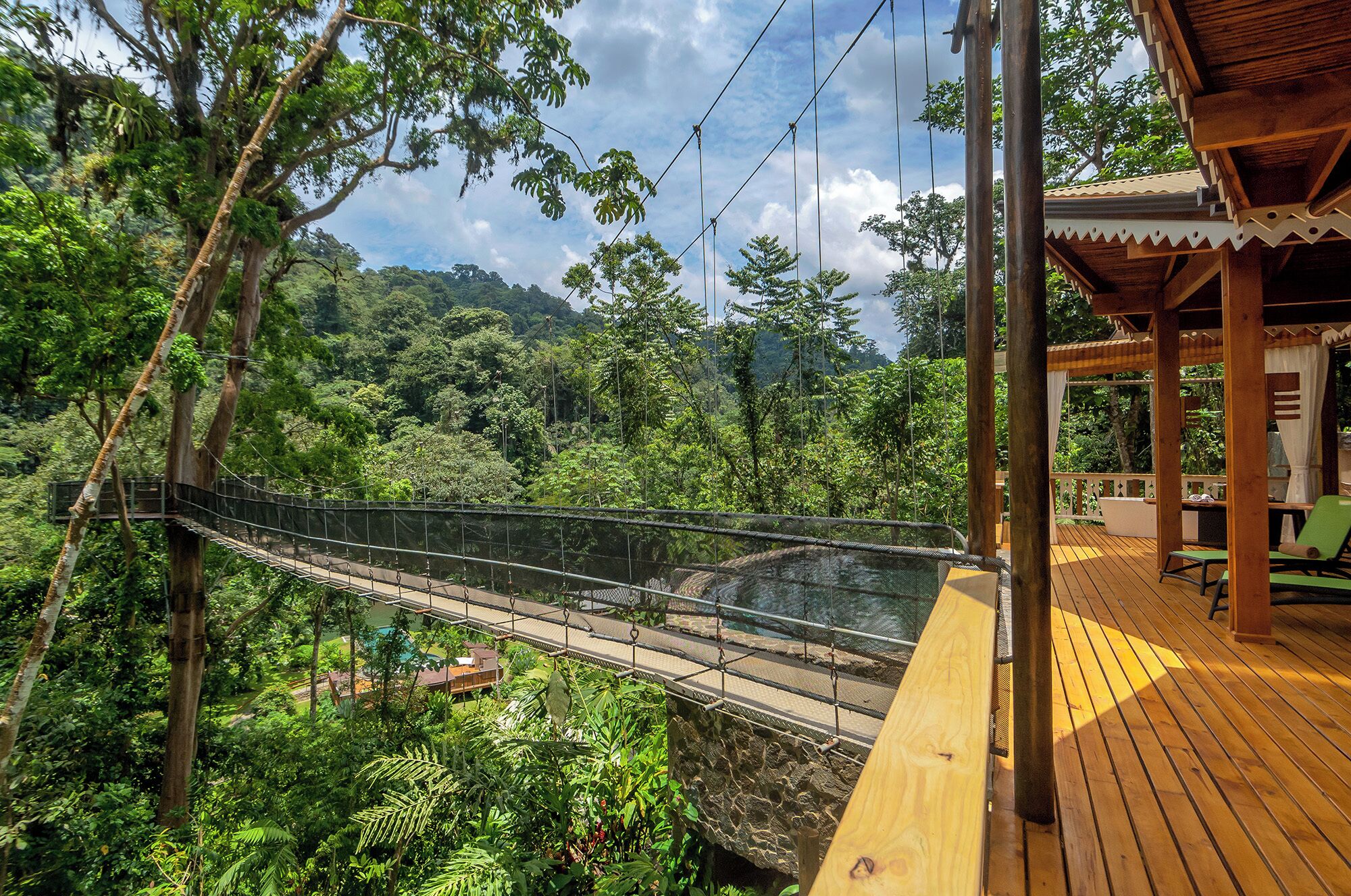 Una terraza de madera con un puente colgante sobre un bosque frondoso, que conduce a un espacio cubierto con asientos.