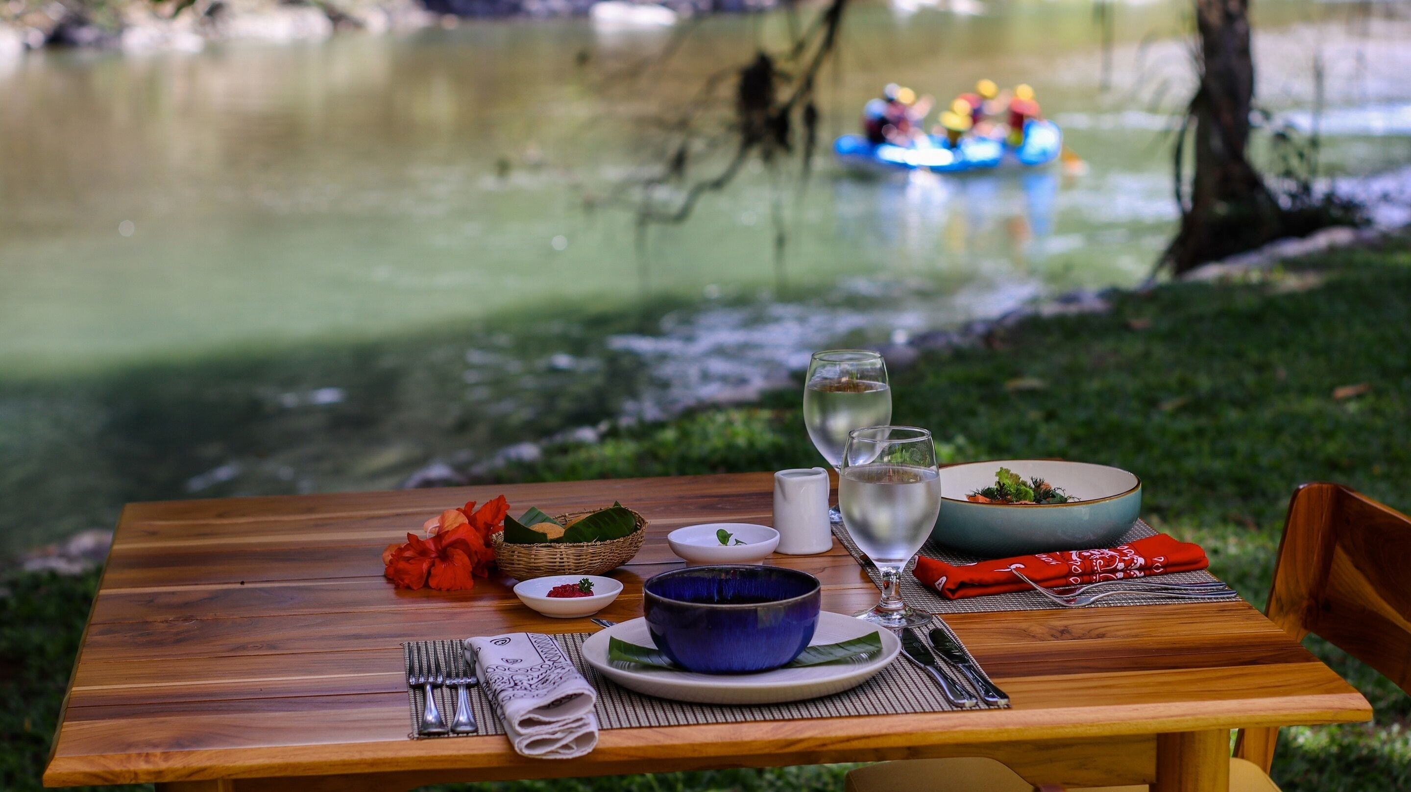A wooden table set with a blue bowl, white plates, silverware, and a red napkin. A wicker basket with red flowers and a white bowl with a green leaf are also visible.