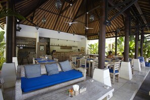 Un área de estar al aire libre con techo, que incluye un banco y mesas de madera, una zona de bar con estantes y botellas, y una estructura de techo de paja.