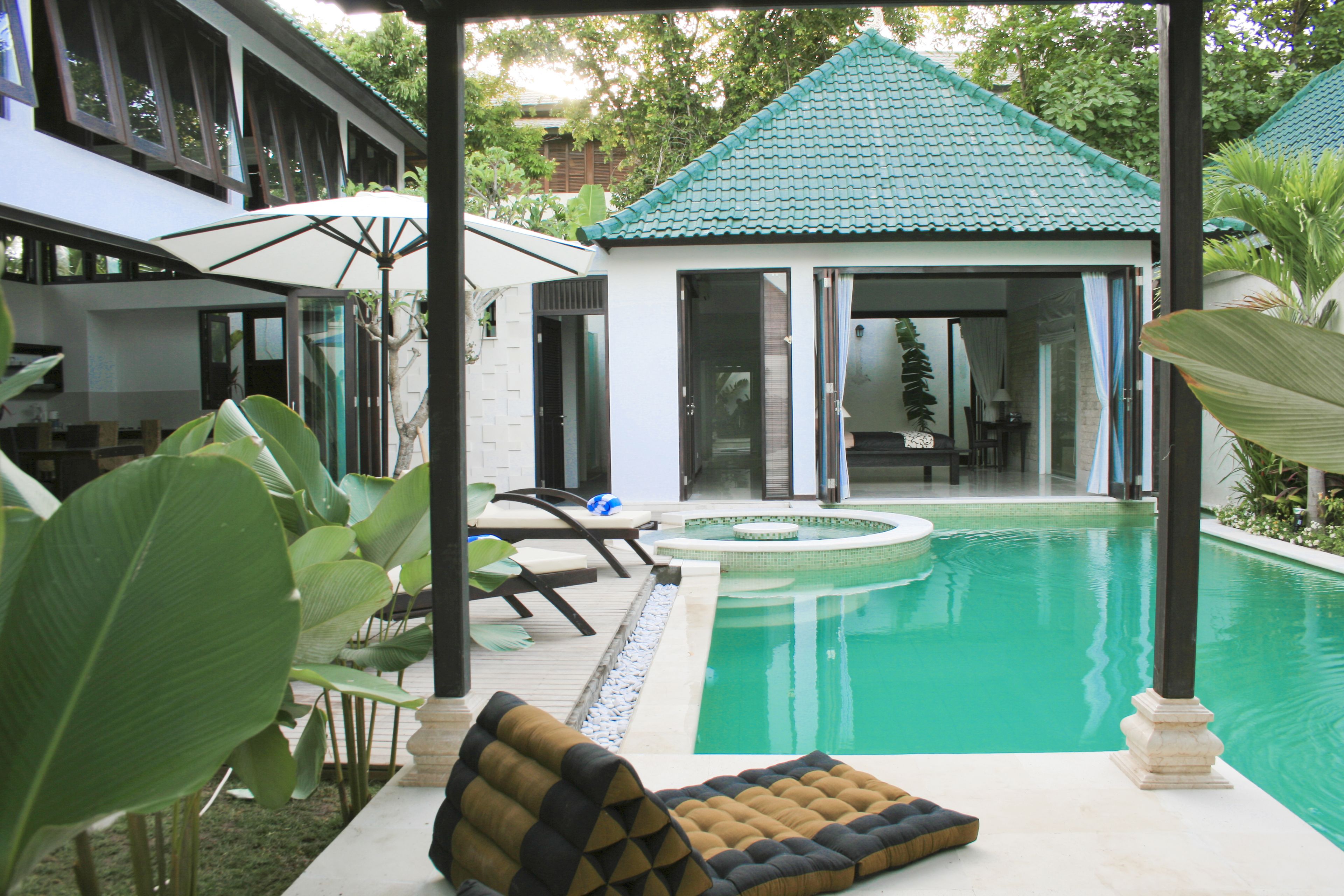 A pool area with lounge chairs, a small building with a tiled roof, and large green plants.