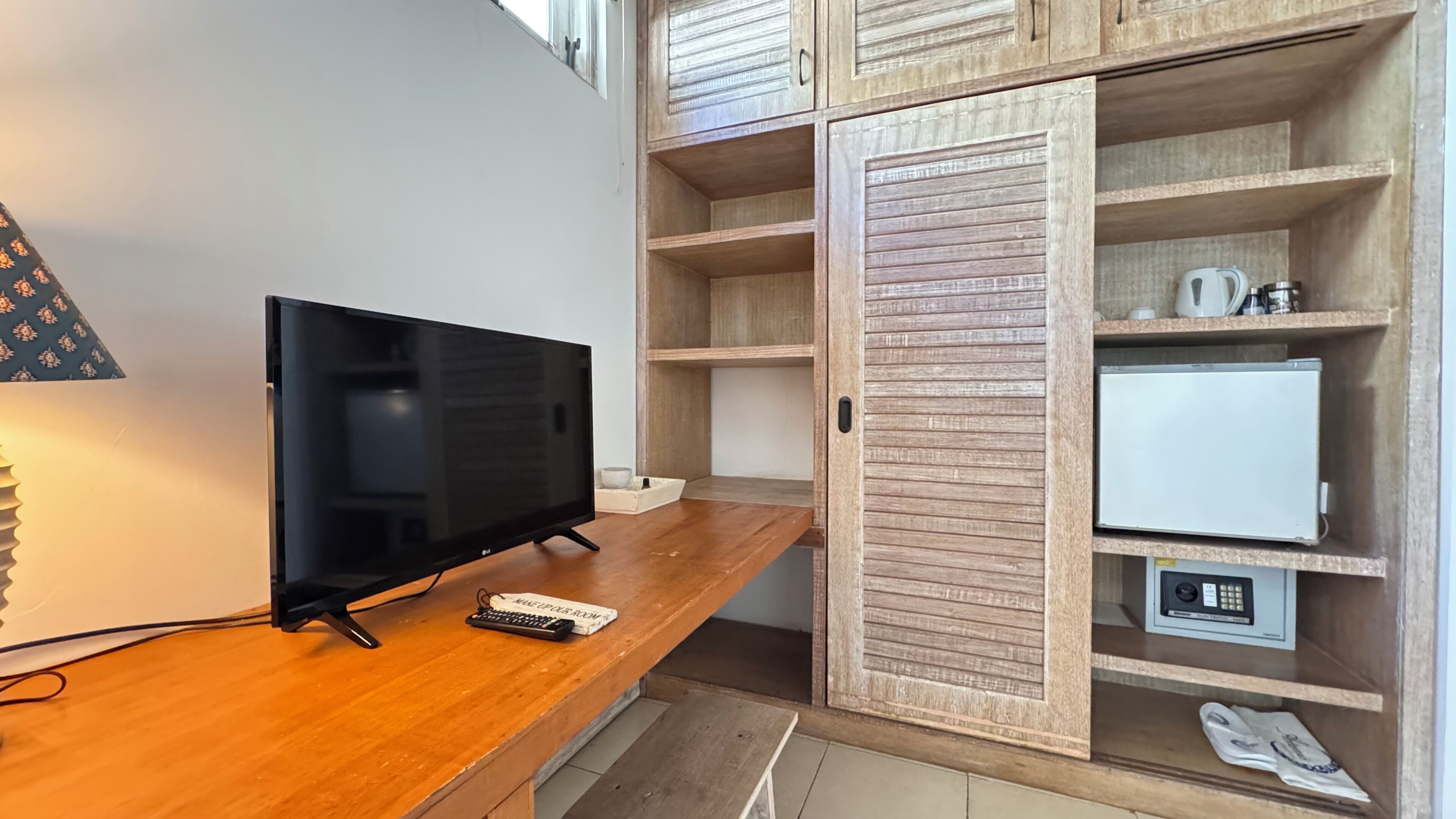 A wooden desk with a television, a remote control, and a keyboard. There is a wardrobe with multiple shelves and a small white appliance.