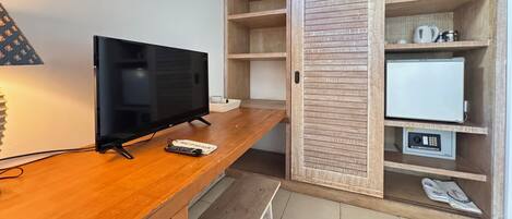 A wooden desk with a television, a remote control, and a keyboard. There is a wardrobe with multiple shelves and a small white appliance.