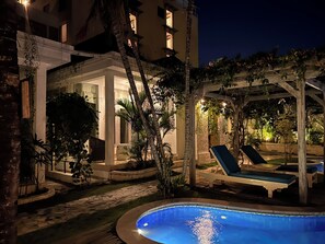 Zona de piscina nocturna con sillones de descanso y una pérgola, rodeada de palmeras y un edificio con ventanas iluminadas.