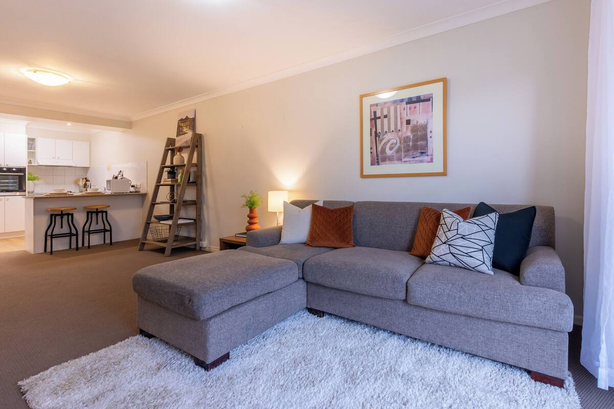 A living room with a grey sectional sofa, a white fluffy rug, and a kitchen in the background.