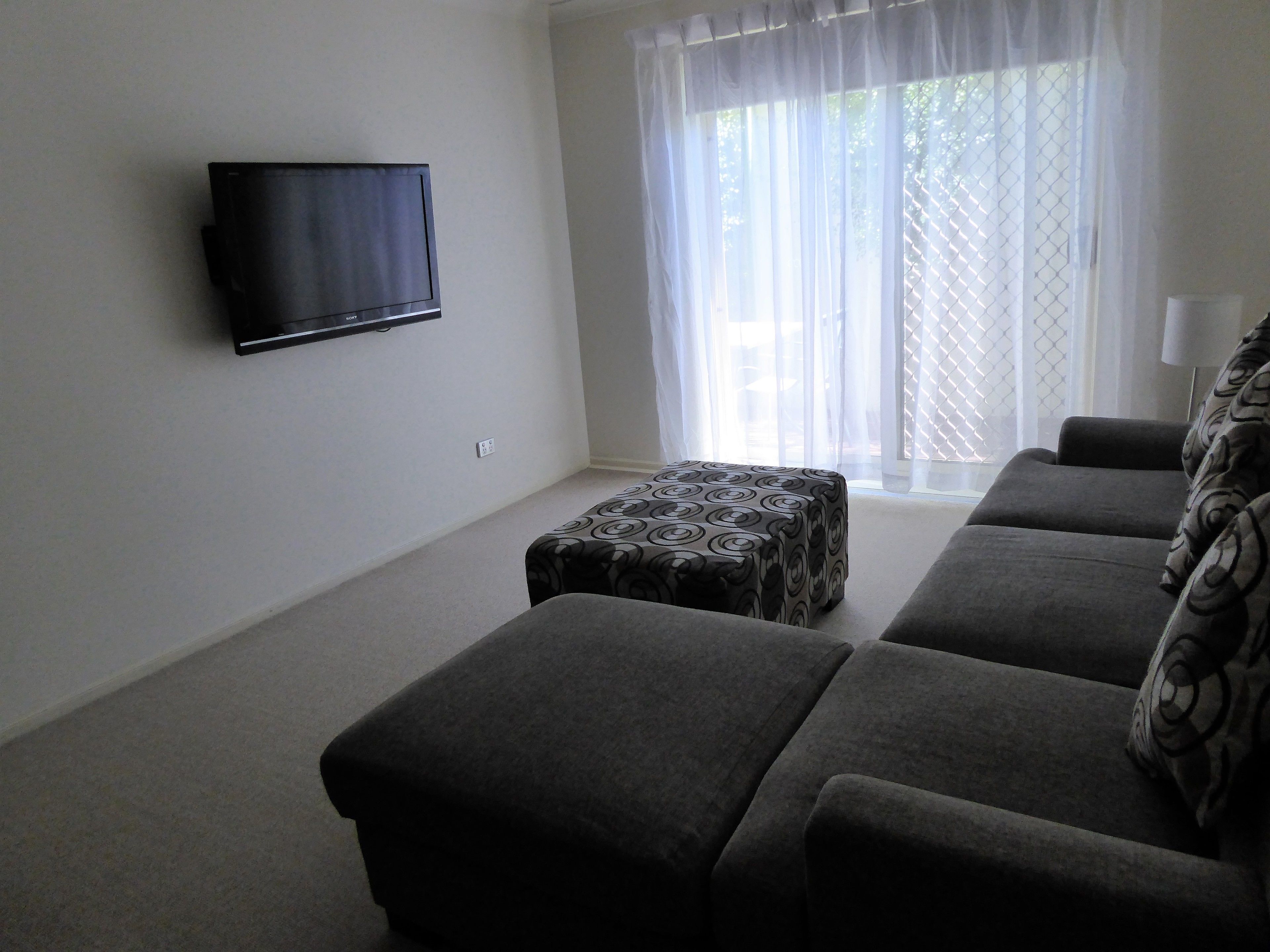 Una sala de estar con un televisor de pantalla plana montado en la pared, un sofá gris y un otomán estampado.