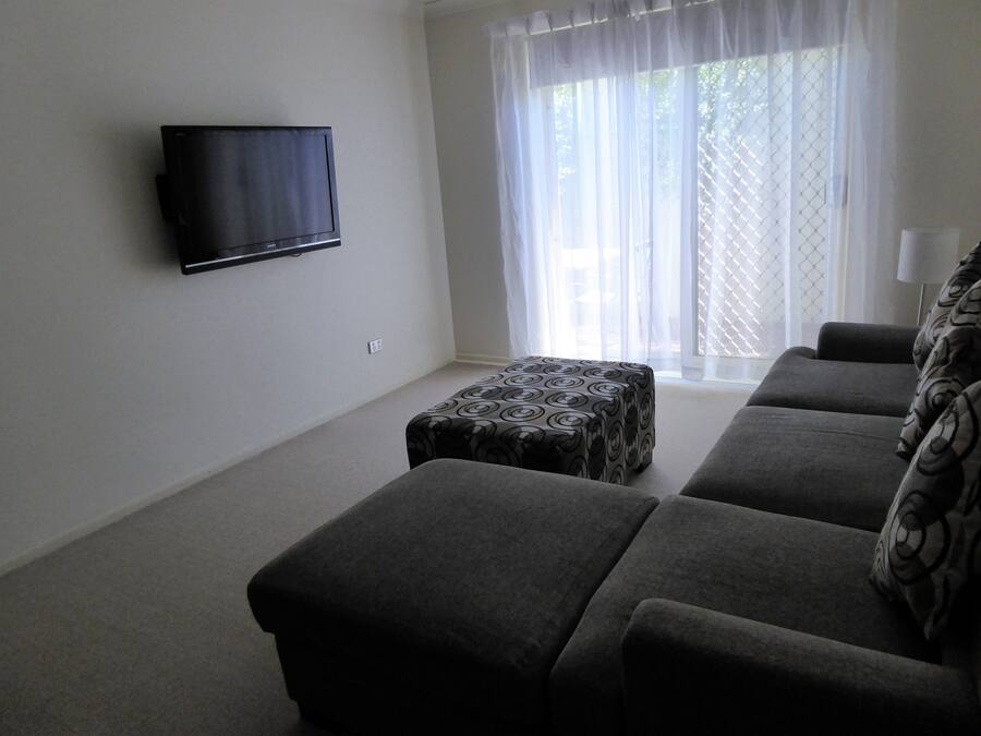 Een woonkamer met een flatscreen televisie aan de muur, een grijze bank en een gestreept voetenbankje.