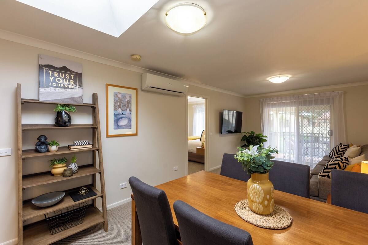 A dining area with a wooden table, chairs, and a decorative shelf with plants and a sign.