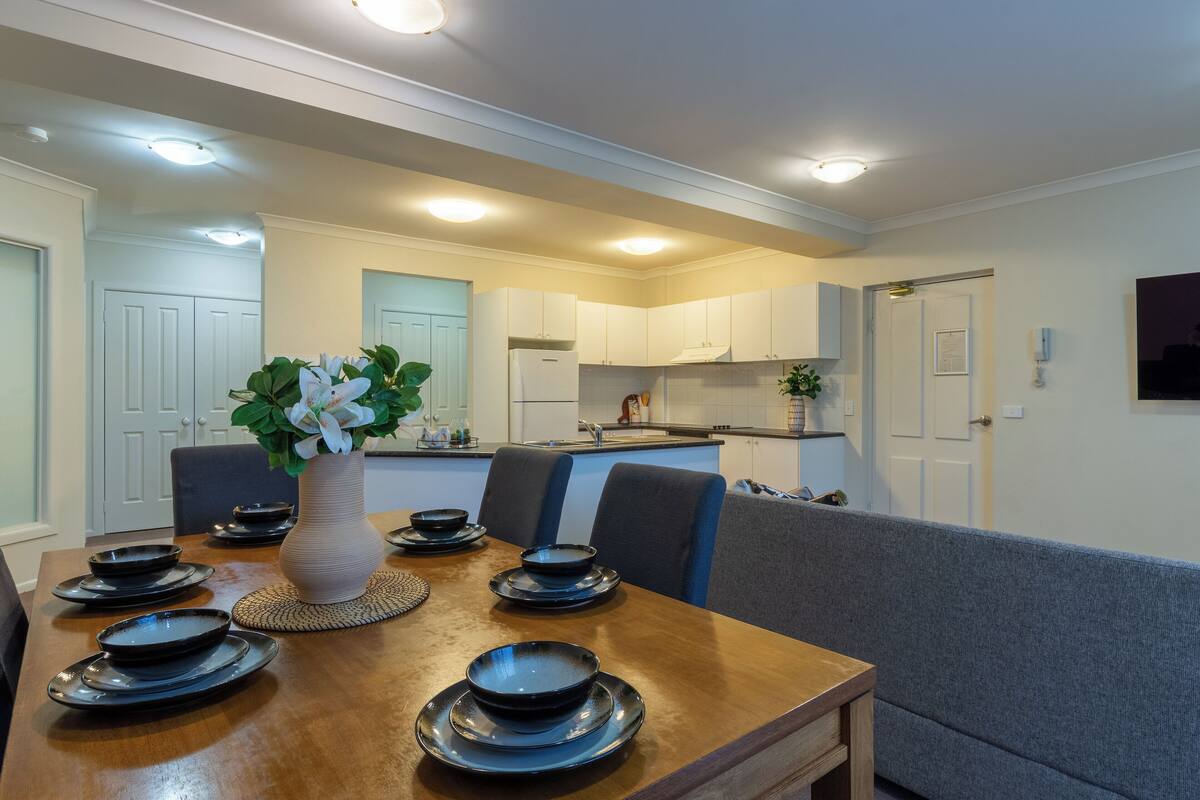A dining area with a wooden table set for four, a vase with white flowers, and a kitchen in the background.