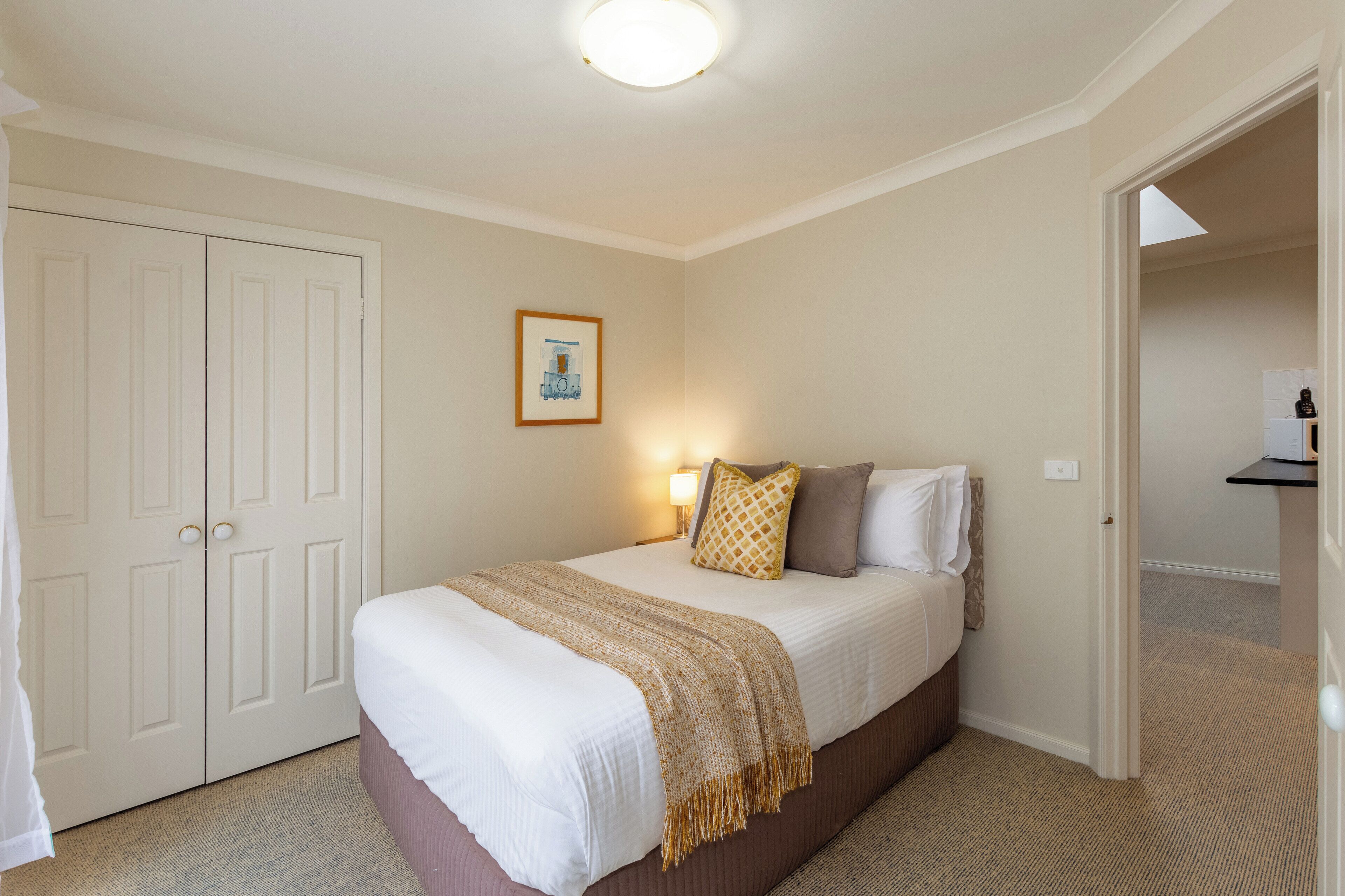 A bedroom with a bed, a bedside table, a lamp, a framed picture on the wall, and a door leading to another room.