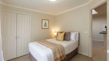 A bedroom with a bed, a bedside table, a lamp, a framed picture on the wall, and a door leading to another room.