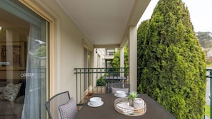 Een balkon met een eettafel gedekt voor twee personen, een bloempot en een hoge, weelderige groene haag.