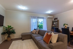 A living room with a sectional sofa, a dining table with chairs, a potted plant, and a window with curtains.