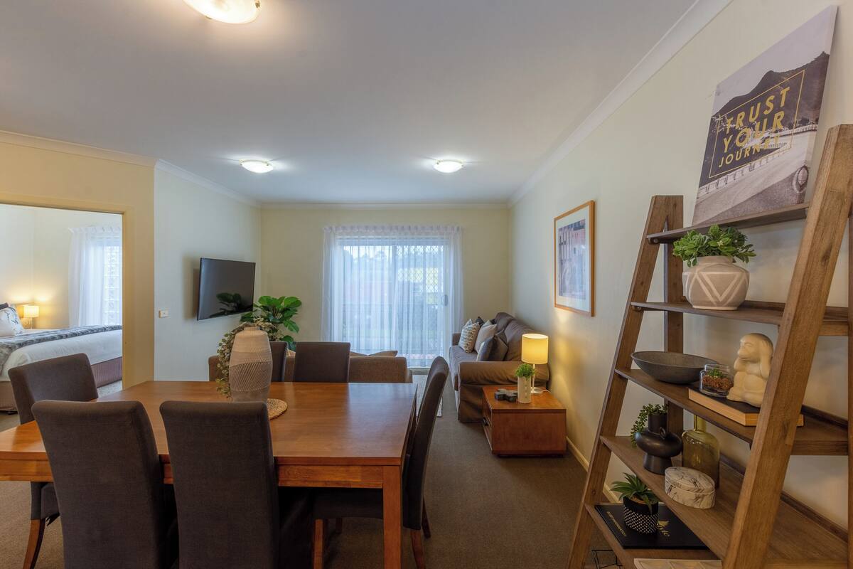 A modern living room with a dining area, a sofa set, a TV mounted on the wall, and a bookshelf with decorative items.
