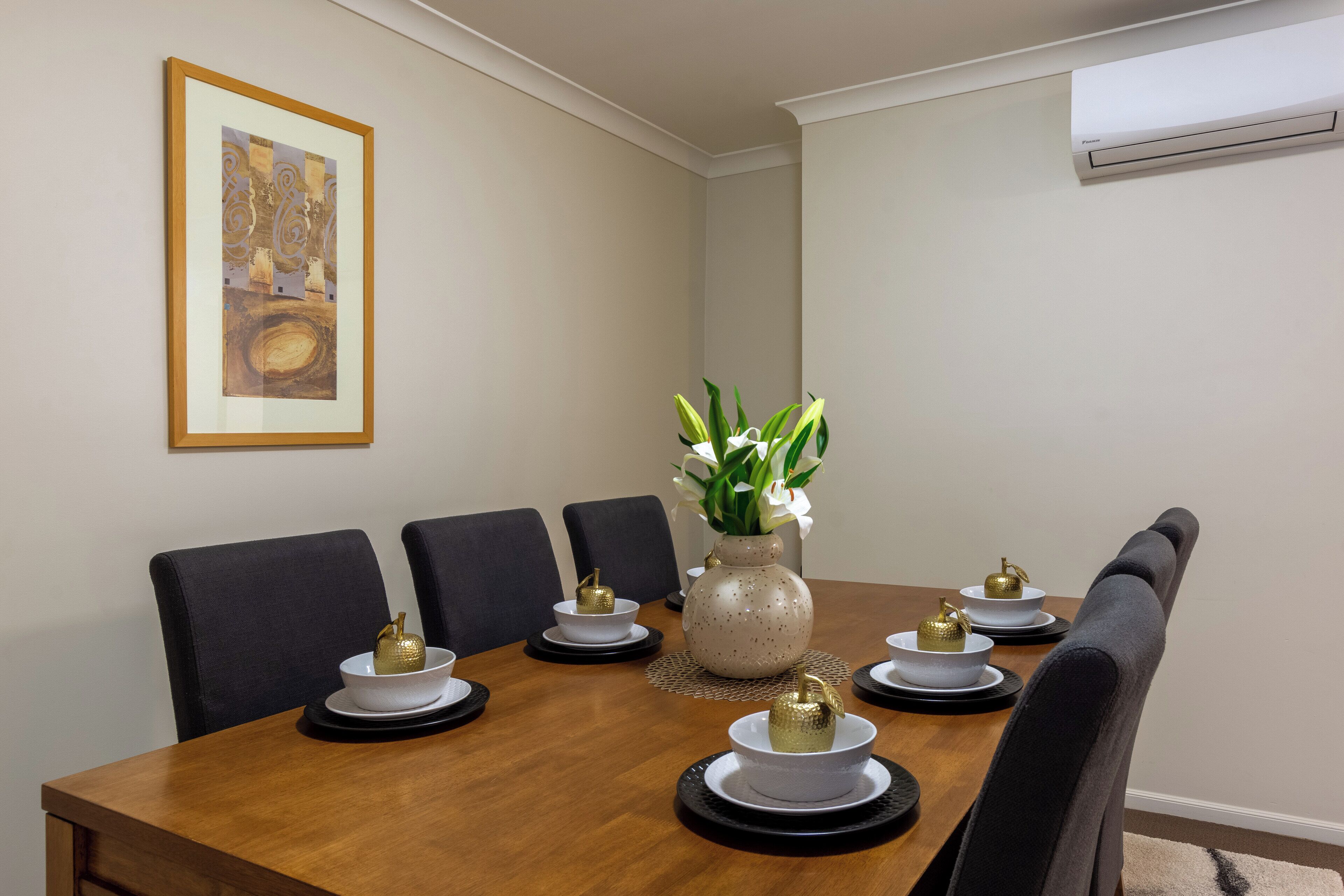 A dining room with a wooden table, black chairs, white plates, gold bowls, and a vase with white flowers.