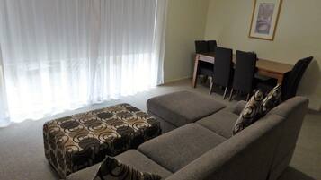 A living room with a sofa set, a dining table with chairs, and a window with sheer curtains.
