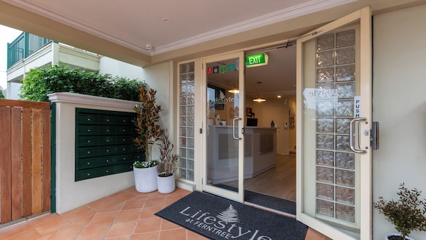 Een hotelentree met een glazen deur, een groen vluchtroutebord, een deurmat met de tekst 'Lifestyle at Ferntree' en een potplant naast de deur.