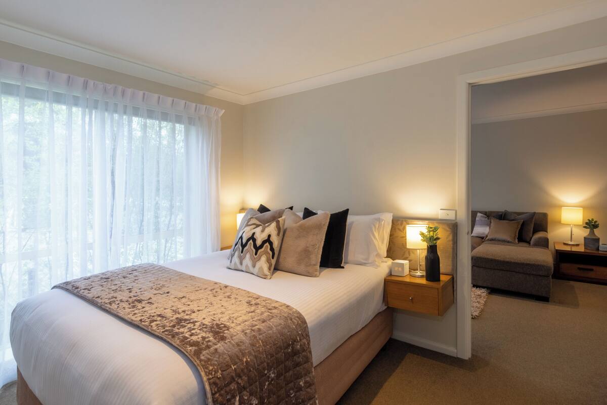 A bedroom with a large bed, bedside tables with lamps, a small sofa, and a view through an open door.