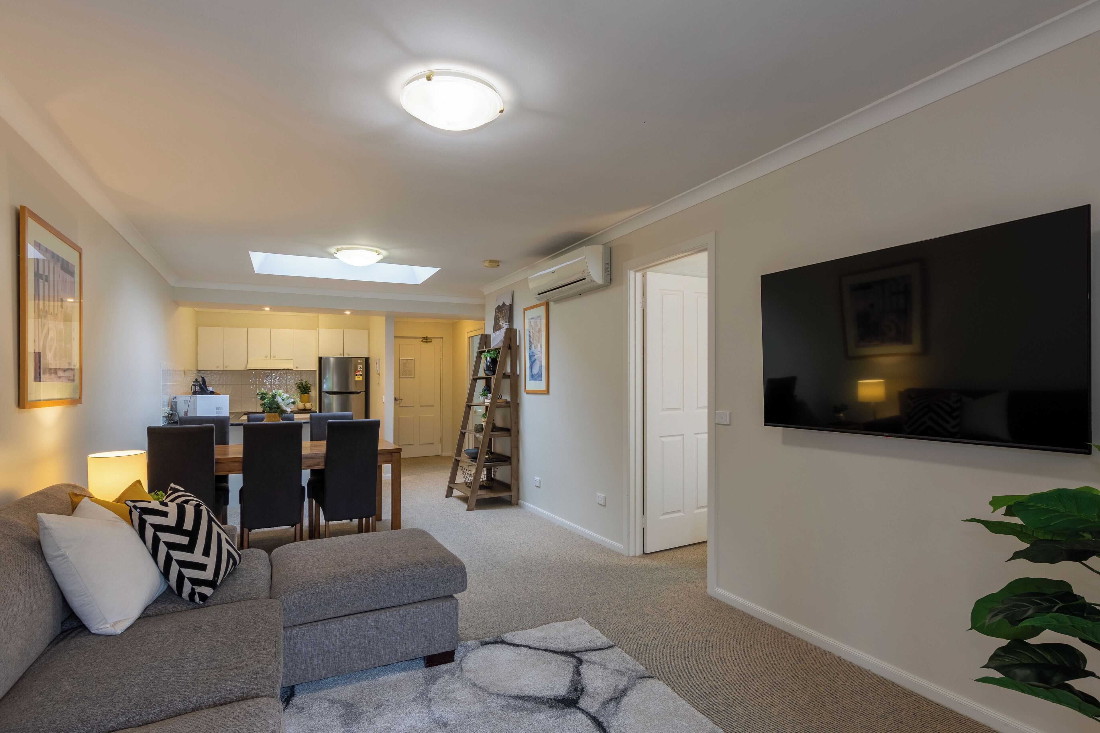 A modern living room with a grey sofa, a flat-screen TV mounted on the wall, and a skylight above the kitchen.