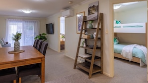 Een kleine leefruimte met een eettafel, stoelen, een stapelbed, een houten ladderrek en een aan de muur bevestigde televisie.