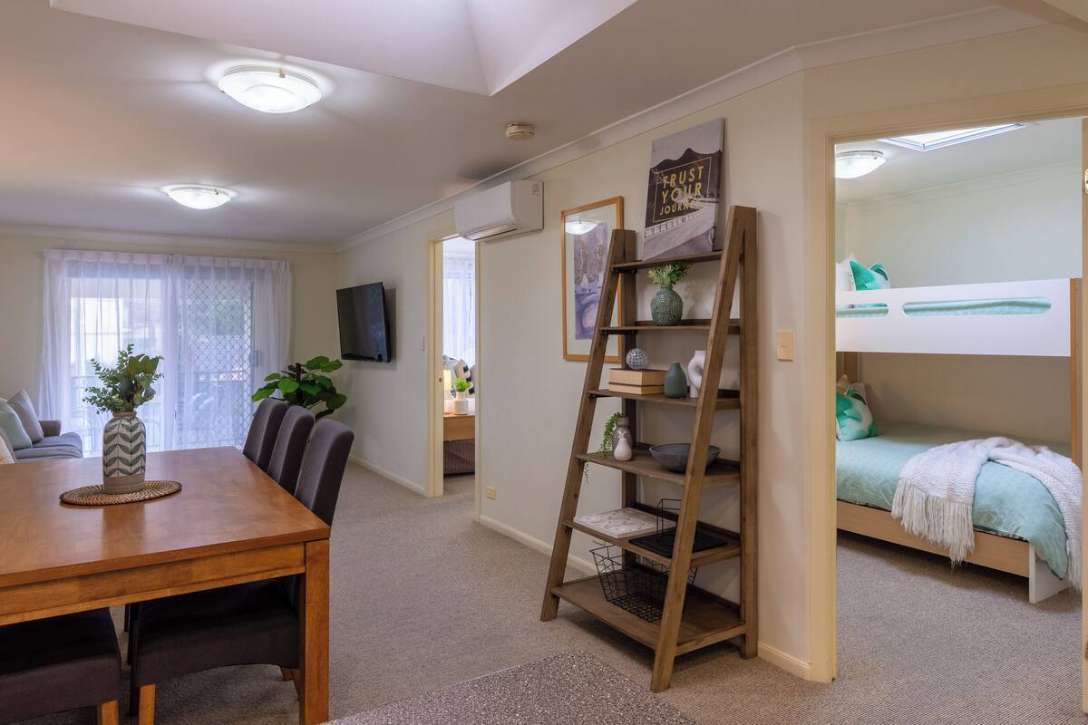 A compact living space with a dining table, chairs, a bunk bed, a wooden ladder shelf, and a TV mounted on the wall.