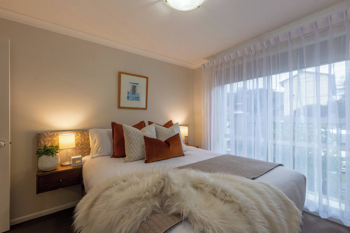A hotel room with a large bed, a nightstand with a lamp, a framed picture on the wall, and sheer curtains on the window.