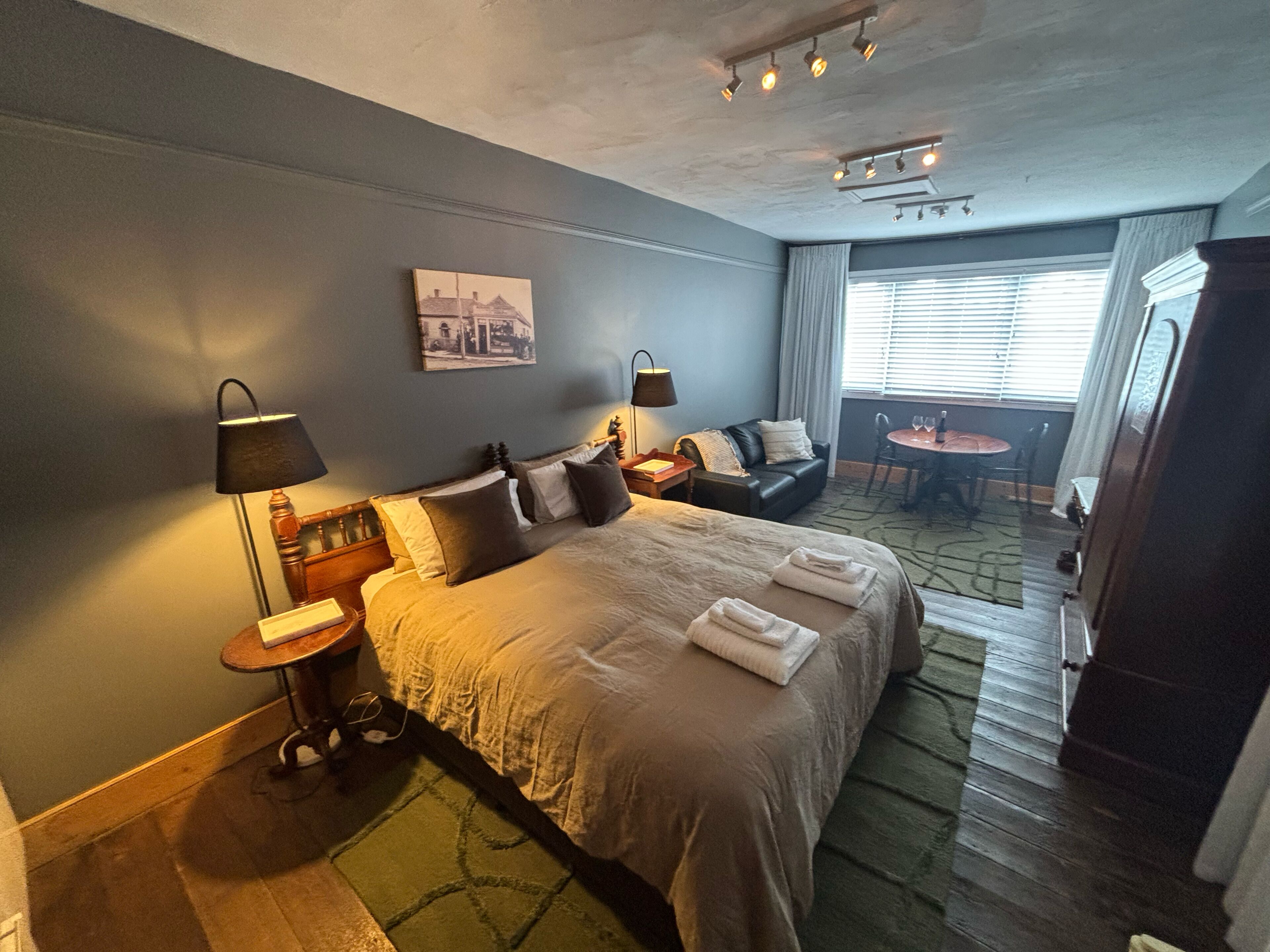 Une chambre d’hôtel avec un lit, des lampes de chevet, une petite table, un canapé, une table à manger et une fenêtre avec des rideaux.