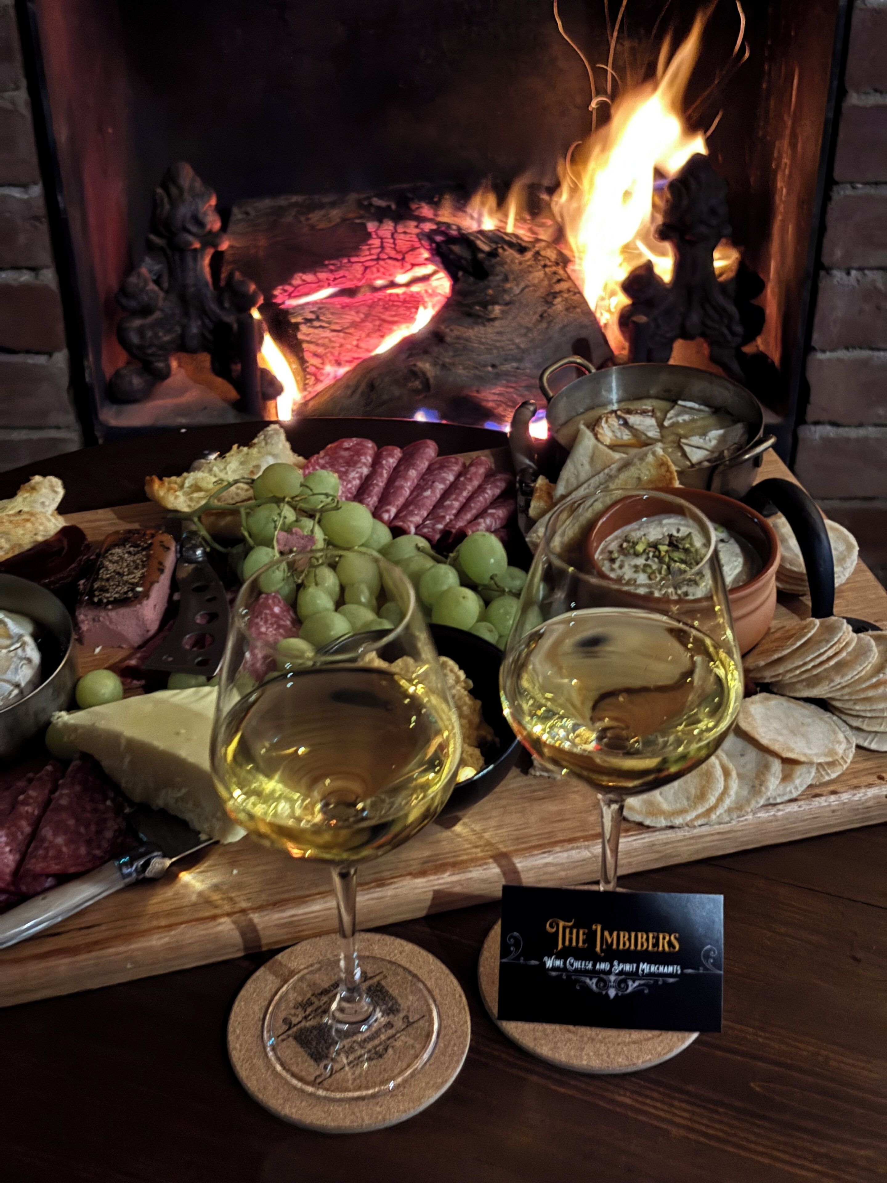 A charcuterie board with various meats, cheeses, and grapes, accompanied by glasses of white wine, set against a backdrop of a fireplace with flames.