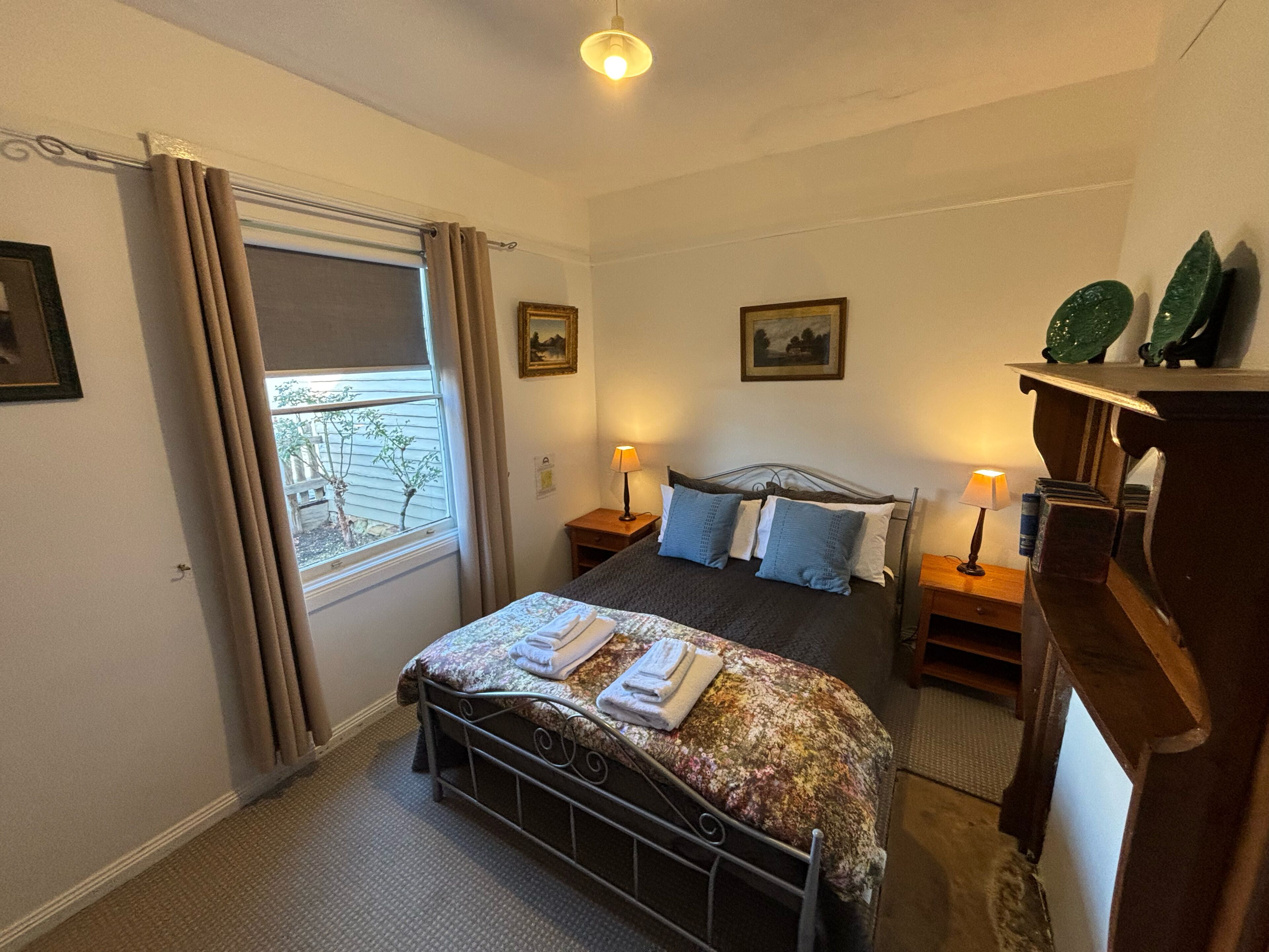 Een slaapkamer met een bed, twee nachtkastjes met lampen, een raam met gordijnen en een kleine plant op de vensterbank.