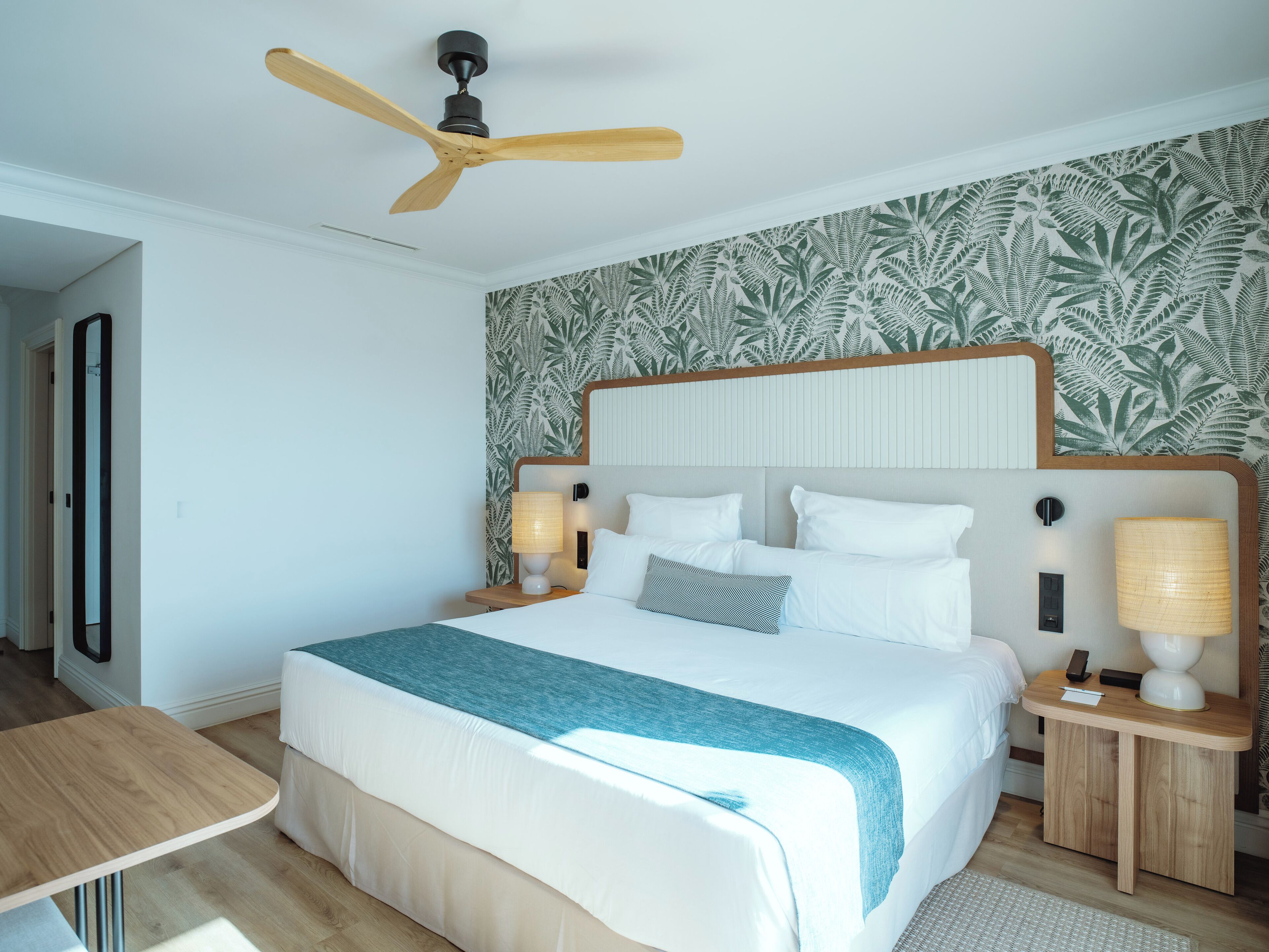 Une chambre d’hôtel avec un grand lit, un ventilateur de plafond, une table de chevet en bois avec une lampe, et un mur orné d’un motif végétal.