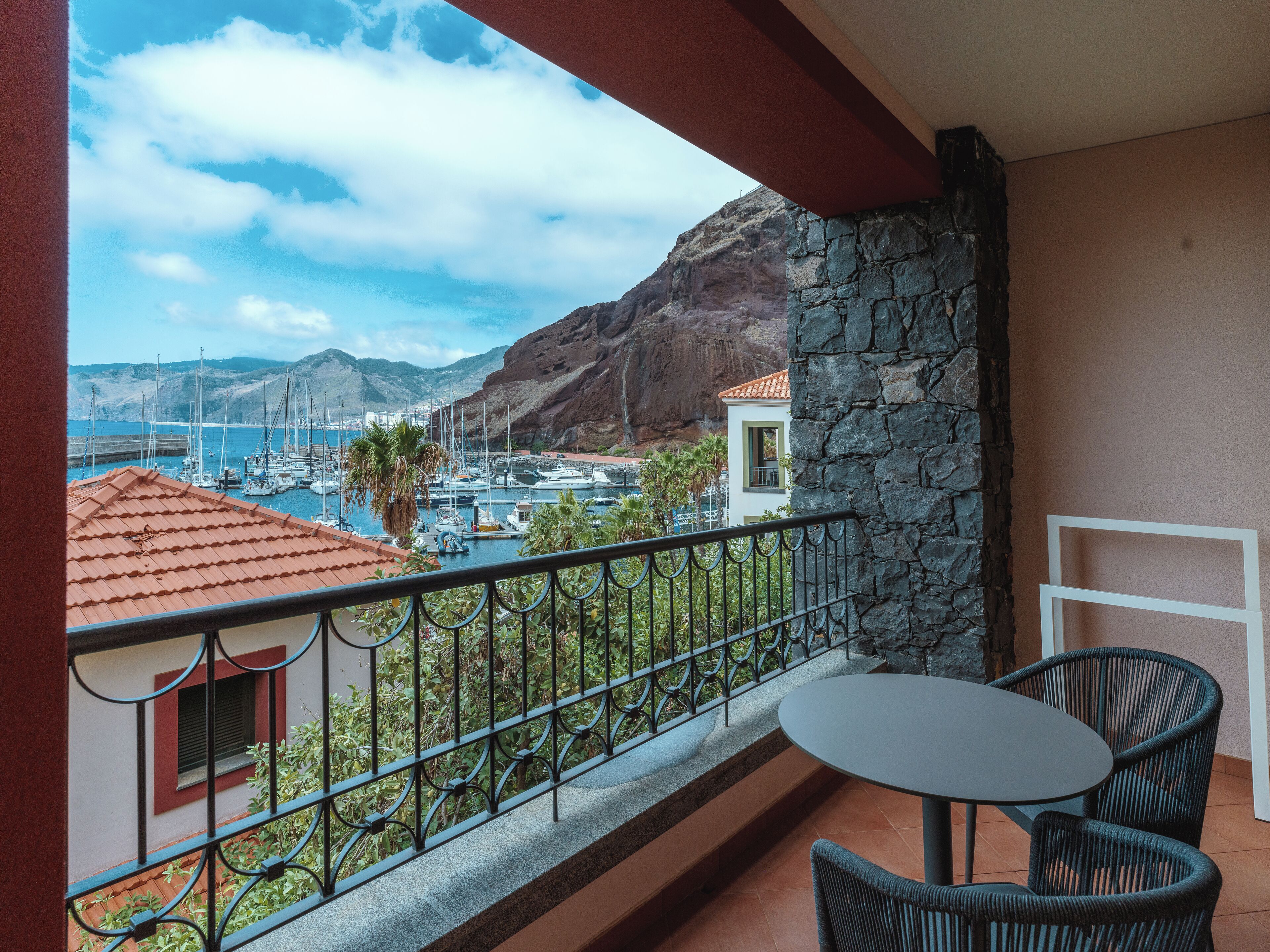 Un balcón con vista a un puerto deportivo, paredes de piedra y una mesa con sillas.