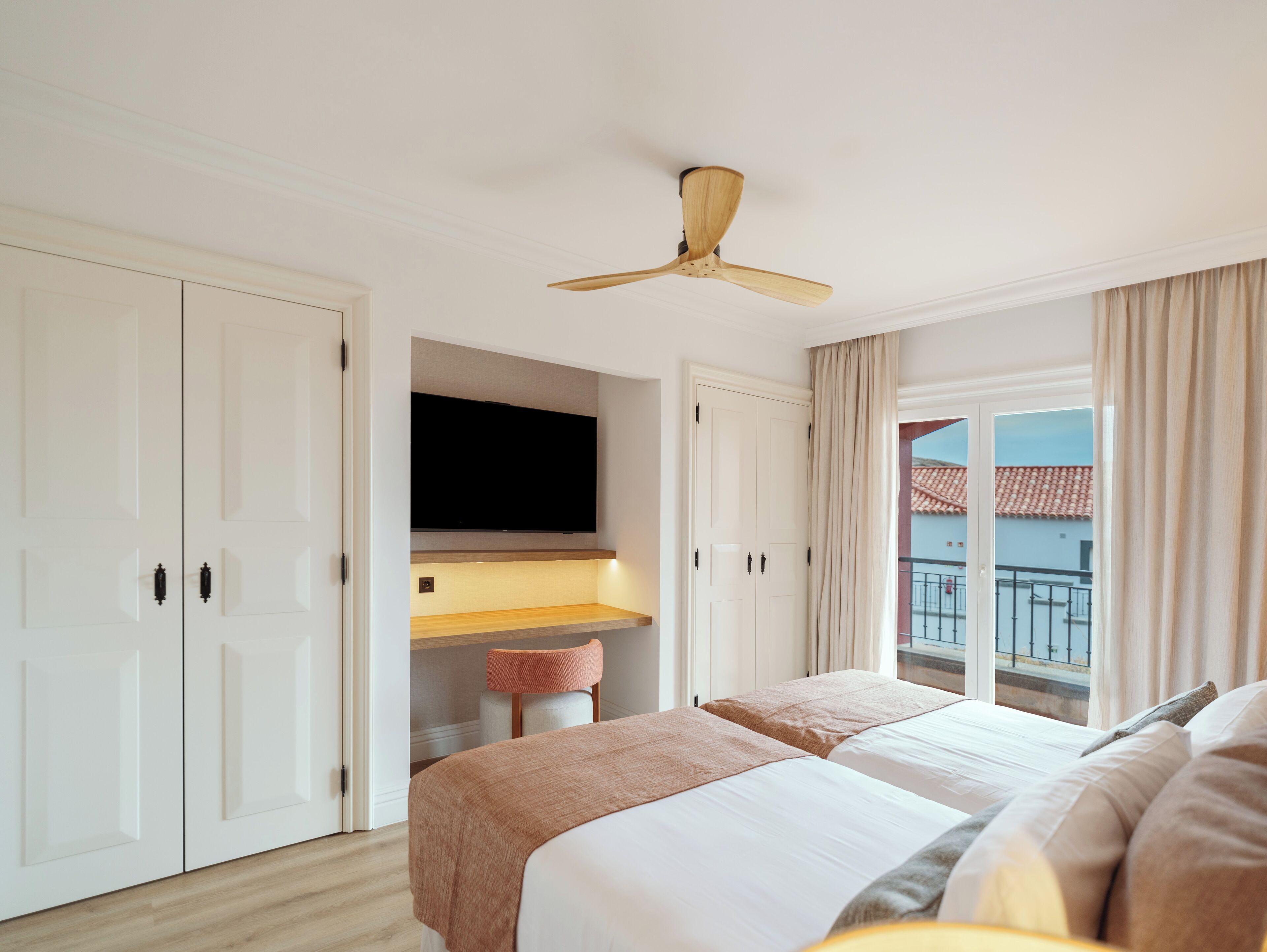 A hotel room with a bed, a desk, a ceiling fan, a TV, and a window with a view of buildings.