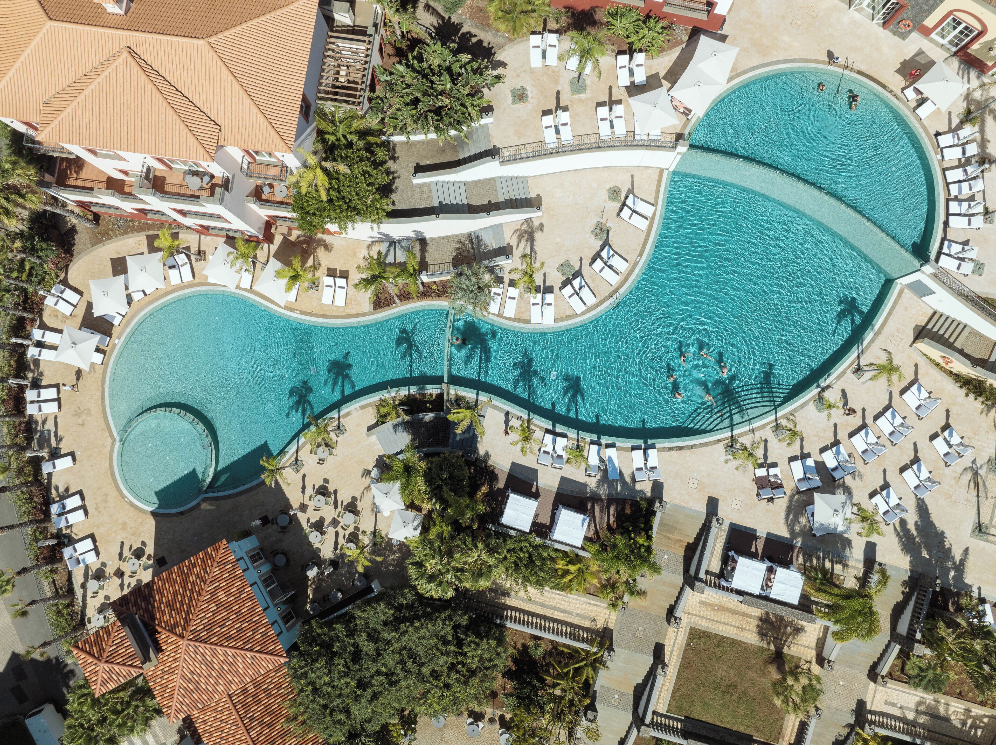 An aerial view of a resort with a large, winding pool surrounded by lounge chairs and umbrellas, palm trees, and a covered area with a tiled roof.