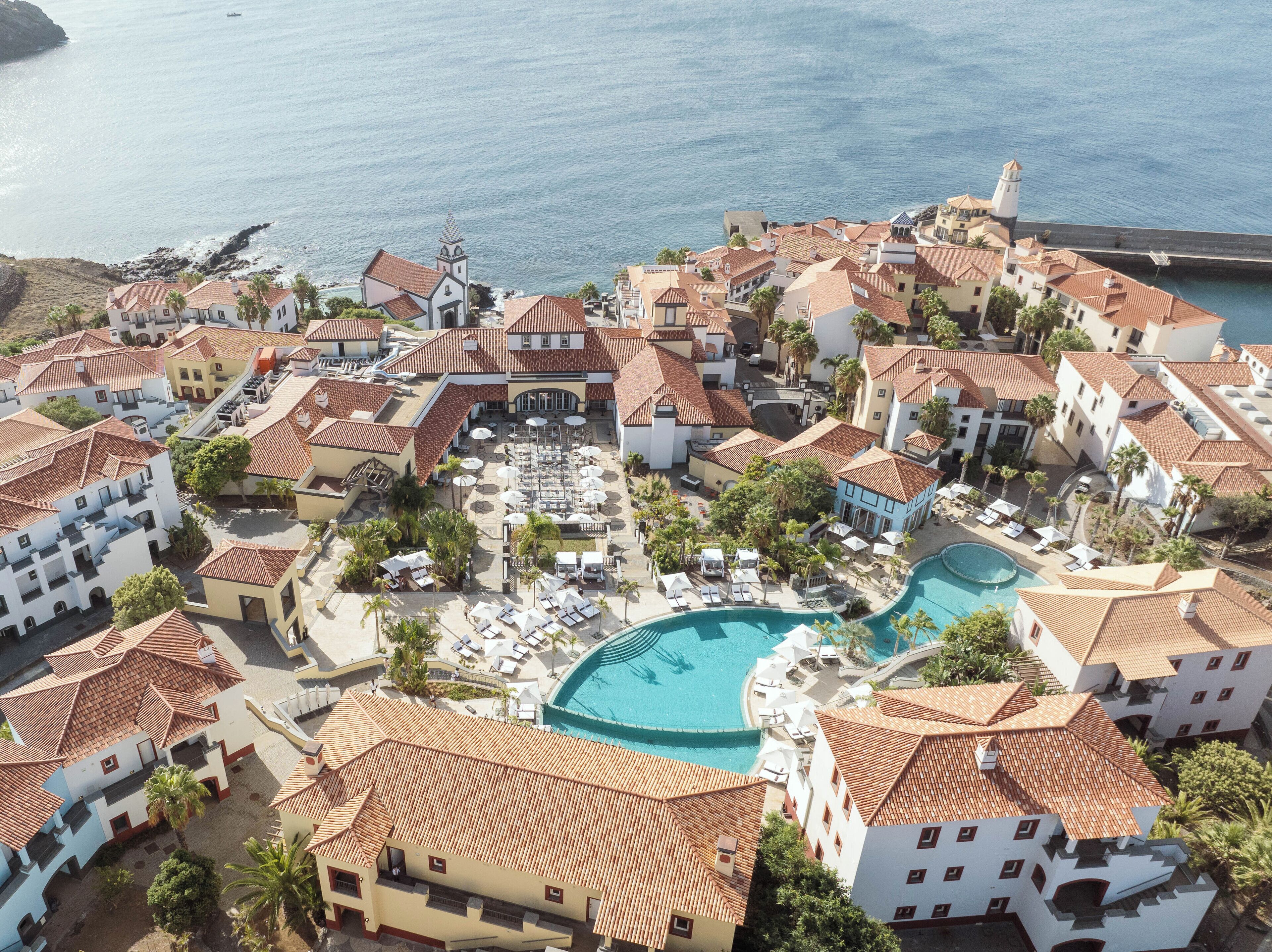 A coastal resort with multiple buildings, a central swimming pool, and a view of the sea.