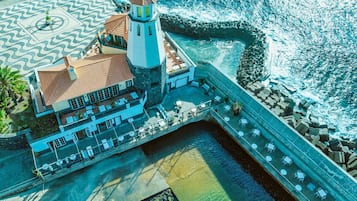 An aerial view of a coastal building with a lighthouse, a terrace with tables and chairs, and a clear view of the sea.