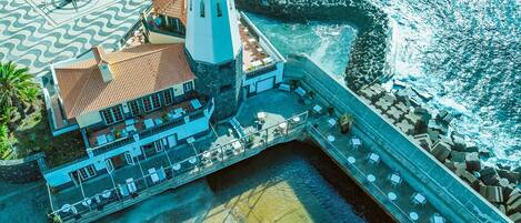 An aerial view of a coastal building with a lighthouse, a terrace with tables and chairs, and a clear view of the sea.