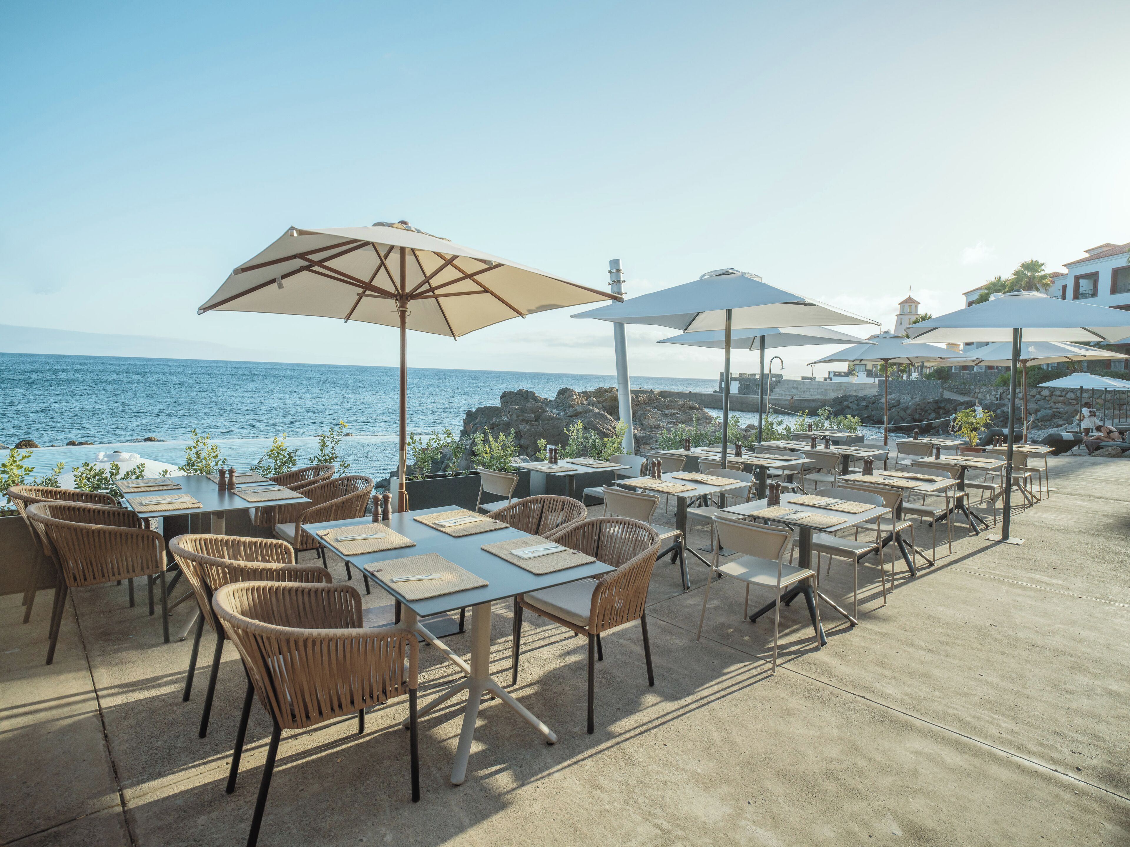 An outdoor dining area with tables and chairs, umbrellas, and a view of the ocean.