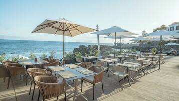 An outdoor dining area with tables and chairs, umbrellas, and a view of the ocean.