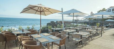 An outdoor dining area with tables and chairs, umbrellas, and a view of the ocean.